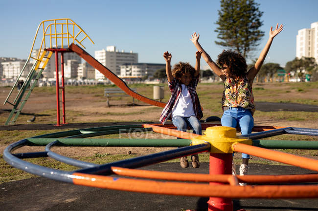 Front view of a young mixed race woman and her pre-teen son enjoying time together playing at a playground, sitting on a carousel with their arms in the air on a sunny day with a swing and buildings in the background — Stock Photo