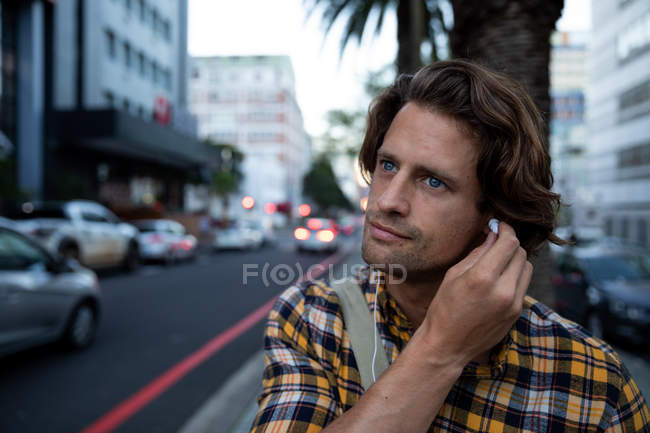 Vista frontal de cerca de un joven caucásico que se pone auriculares en una calle urbana concurrida durante su viaje nocturno - foto de stock