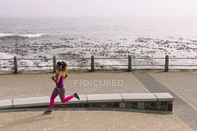 Vista lateral de una joven mujer caucásica con ropa deportiva corriendo durante un entrenamiento en un día soleado junto al mar - foto de stock