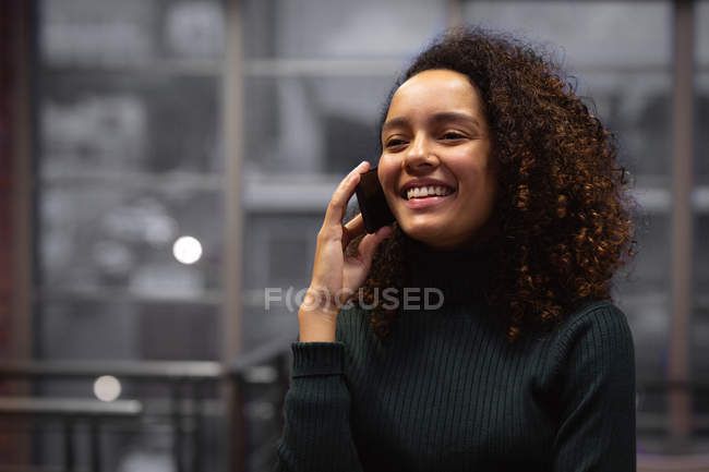 Nahaufnahme einer lächelnden jungen Frau mit gemischter Rasse, die im Büro eines kreativen Unternehmens auf einem Smartphone steht und spricht — Stockfoto