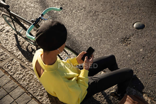 HIgh ângulo vista de um jovem elegante mestiço transgênero adulto na rua, mensagens de texto no smartphone vestindo uma boina com uma bicicleta no fundo — Fotografia de Stock