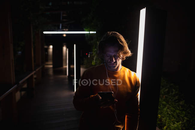 Vista frontale di un giovane caucasico in piedi in una strada di notte ad ascoltare musica con gli auricolari, guardando uno smartphone — Foto stock