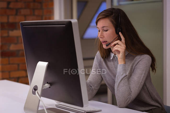Vista frontal de cerca de una joven mujer caucásica sentada en un escritorio trabajando en la oficina de un negocio creativo usando una computadora y hablando con un auricular telefónico - foto de stock