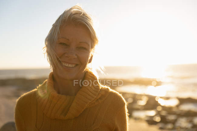 Porträt einer reifen kaukasischen Frau, die bei Sonnenuntergang am Meer in die Kamera lächelt — Stockfoto