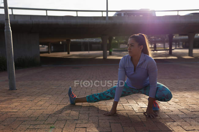 Vista frontal de cerca de una joven mujer caucásica vestida con ropa deportiva sentada y tocando el suelo con una pierna estirada mientras hace ejercicio en un parque, retroiluminada por la luz del sol - foto de stock