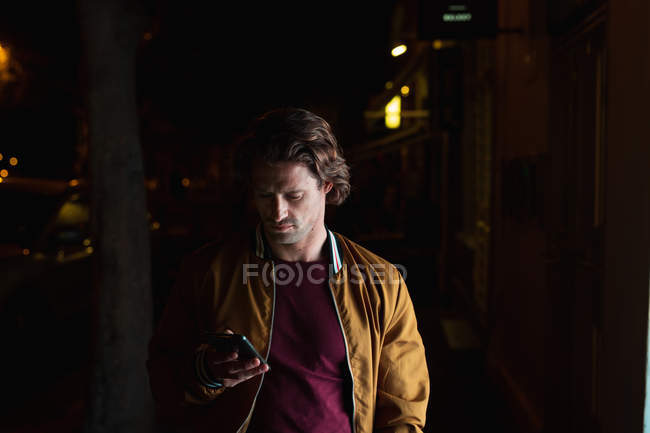 Frontansicht eines jungen kaukasischen Mannes, der nachts mit einem Smartphone auf einer Straße steht — Stockfoto