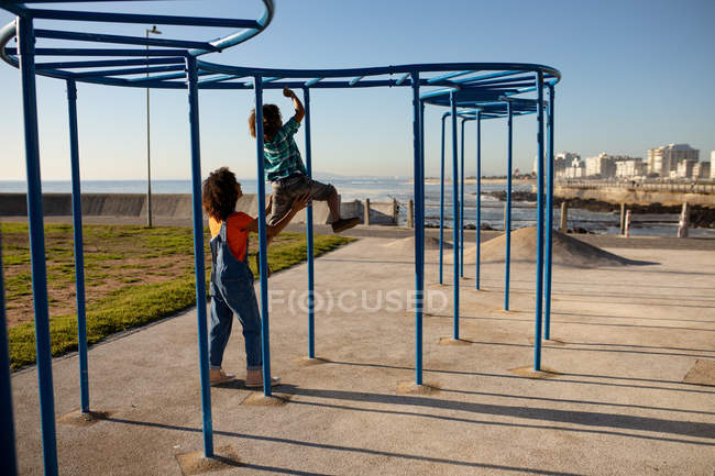 Vista trasera de una joven mestiza y su hijo preadolescente disfrutando del tiempo juntos jugando en un parque infantil junto al mar, bajo un marco de escalada en un día soleado - foto de stock