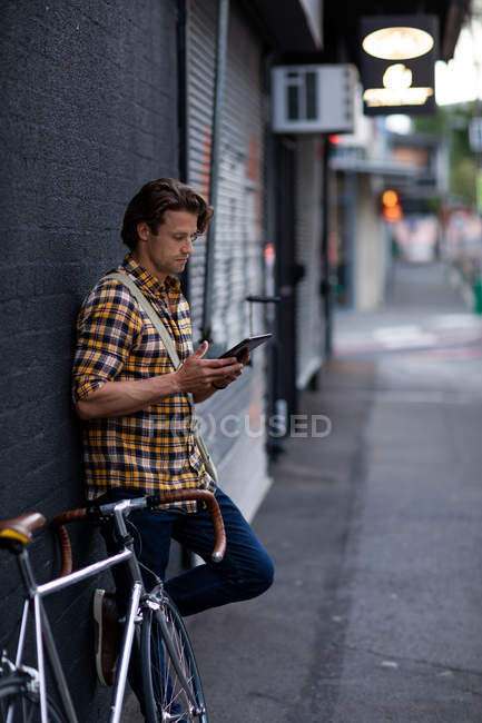 Vue latérale d'un jeune homme caucasien tenant une tablette avec un vélo à côté de lui appuyé contre un mur dans une rue urbaine pendant son trajet du soir à la maison — Photo de stock