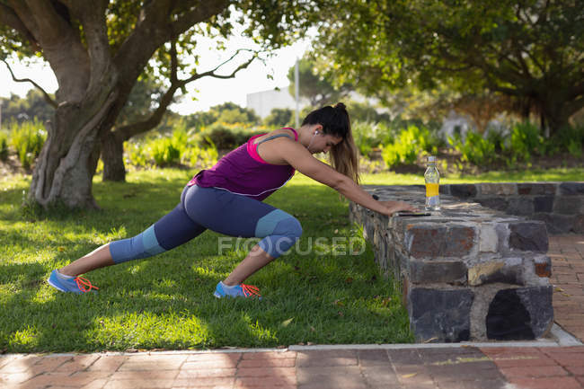 Vista laterale di una giovane donna caucasica che indossa vestiti sportivi spingendo contro un muro e allungandosi durante un allenamento in un parco — Foto stock