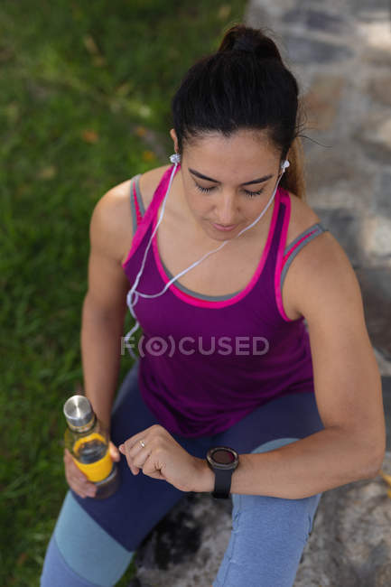 Высокий вид на молодую кавказскую женщину в спортивной одежде, держащую бутылку воды, проверяющую свои умные часы и слушающую музыку в наушниках во время тренировки в солнечный день в парке — стоковое фото