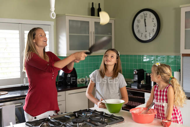 Front view of a young Caucasian pregnant woman making pancakes in the kitchen at home with her tween and younger daughter — стокове фото