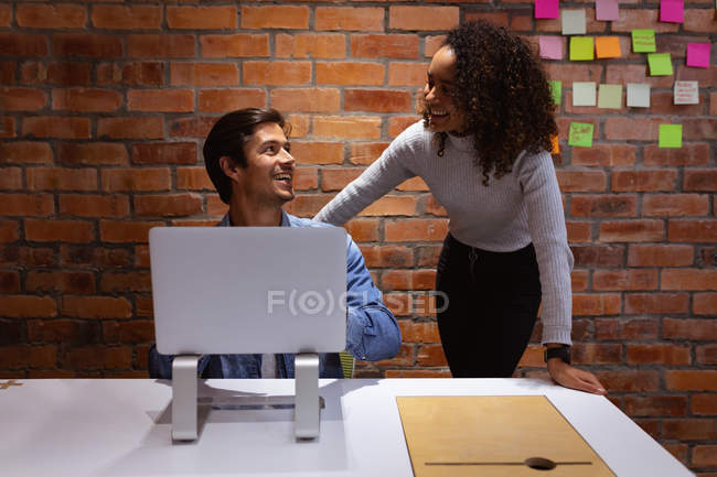 Vista frontal de cerca de una joven mujer de raza mixta de pie y un joven hombre caucásico sentado usando una computadora portátil sonriendo y hablando juntos en una mesa mientras trabajan en la oficina de un negocio creativo - foto de stock