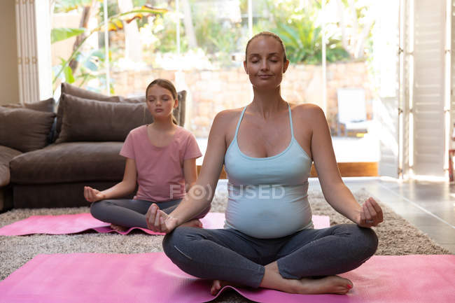 Front view of a young Caucasian pregnant woman doing yoga with her tween daughter in their sitting room — Stock Photo