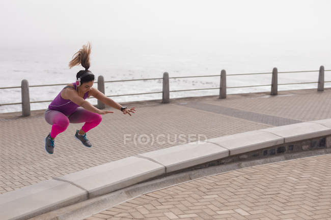 Vista frontale di una giovane donna caucasica che indossa vestiti sportivi saltando su un muro basso durante un allenamento in una giornata di sole in riva al mare — Foto stock