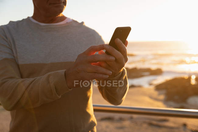 Vista frontal de cerca de un hombre caucásico maduro usando un teléfono inteligente junto al mar al atardecer - foto de stock