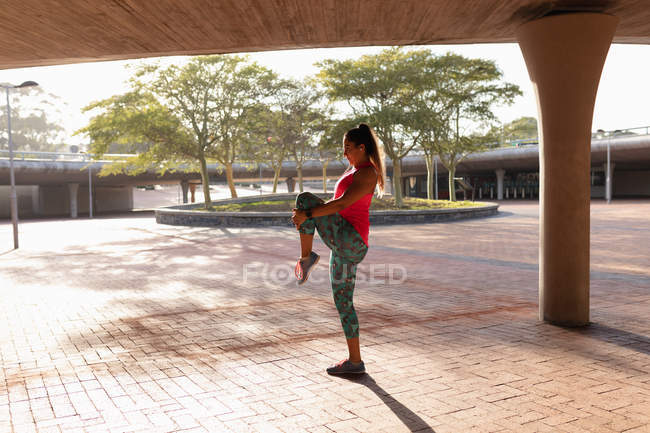 Вид збоку молоді кавказька жінка носить спортивний одяг стоячи і потягнувши її ногу до грудей і розтягування під мостом під час тренування в парку — стокове фото