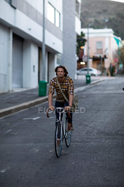 Vista frontal de um jovem caucasiano andando de bicicleta em uma rua urbana, viajando para casa do trabalho à noite — Fotografia de Stock