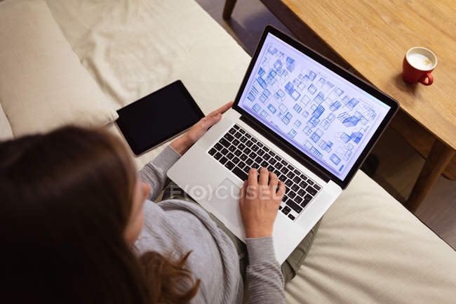 Ansicht einer jungen kaukasischen Frau mit einem Laptop, die auf einem Sofa im Loungebereich eines kreativen Büros sitzt und eine Tasse Kaffee auf dem Tisch vor sich hat — Stockfoto