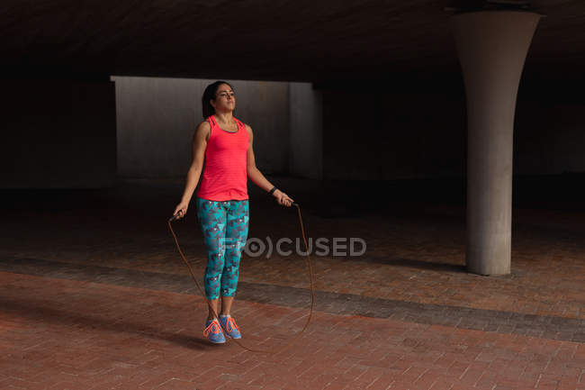 Front view of a young Caucasian woman wearing sports clothes jumping with a skipping rope during a workout under a bridge in a park — Stock Photo
