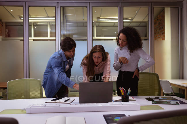 Vue de face d'une jeune femme métissée et d'une jeune femme et homme caucasiens debout autour d'un ordinateur portable travaillant ensemble dans le bureau d'une entreprise créative — Photo de stock