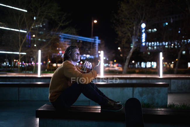 Vista lateral de un joven caucásico sentado en una pared en la calle por la noche hablando por teléfono con auriculares puestos, con un monopatín a su lado - foto de stock