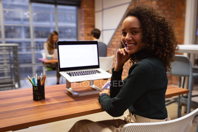 Портрет крупним планом молодої змішаної раси жінки, яка працює в офісі творчого бізнесу, сидячи за столом за допомогою ноутбука, повертаючись і посміхаючись до камери, з колегами, які працюють за столом на задньому плані — стокове фото
