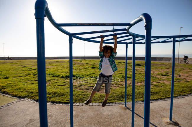 Front view of a mixed race pre-teen boy playing at a playground by the sea, hanging from a climbing frame on a sunny day — Stock Photo