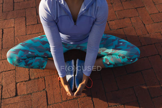 Elevated low section view of woman wearing sports clothes sitting on a path, holding her feet and stretching while working out in a park — Stock Photo