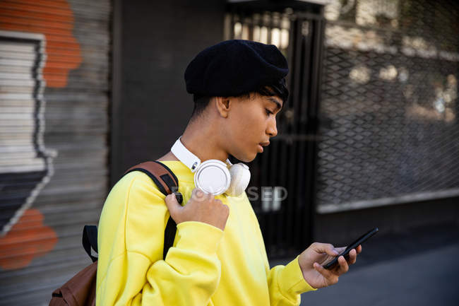 Vue latérale d'un jeune adulte transgenre mixte à la mode dans la rue, à l'aide d'un smartphone, dans un béret et un casque — Photo de stock