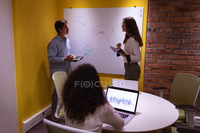 Seitenansicht einer jungen kaukasischen Frau und eines Mannes, die im Büro eines kreativen Unternehmens an einem Whiteboard stehen, Tablet und Laptop in der Hand halten und reden, während eine Kollegin — Stockfoto