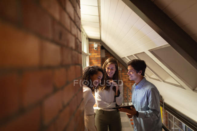 Seitenansicht einer jungen Frau mit gemischter Rasse und einer jungen kaukasischen Frau und einem Mann, die lächelnd im Büro eines kreativen Unternehmens stehen und auf einen Tablet-Computer blicken, den der Mann in der Hand hält — Stockfoto