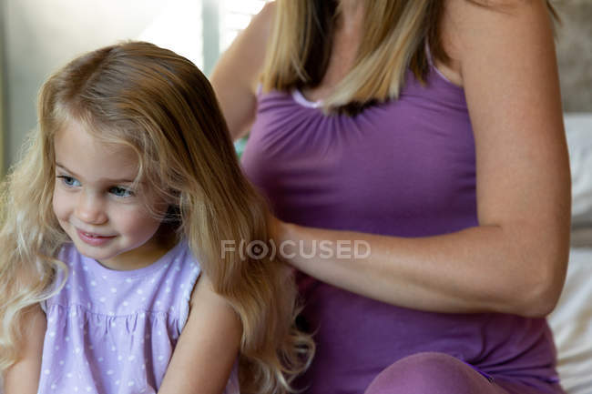 Вид спереди на молодую кавказскую беременную женщину, расчесывающую волосы своей маленькой дочери в своей спальне — стоковое фото