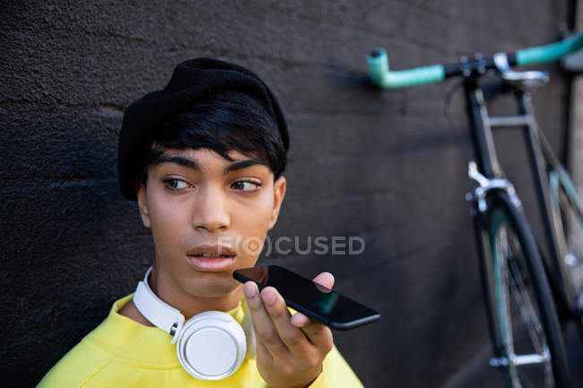 Front view of a fashionable young mixed race transgender adult in the street, talking on the smartphone in a beret and headphones with a bike in a background — Stock Photo