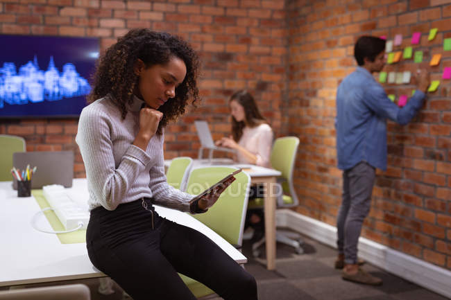 Вид сбоку: молодая женщина смешанной расы, работающая в офисе креативного бизнеса, сидящая на столе за планшетным компьютером, с двумя коллегами, работающими на заднем плане — стоковое фото