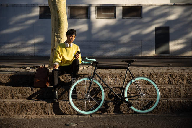 Front view of a fashionable young mixed race transgender adult in the street, holding a smartphone and a coffee cup sitting on steps next to a bike — Stock Photo