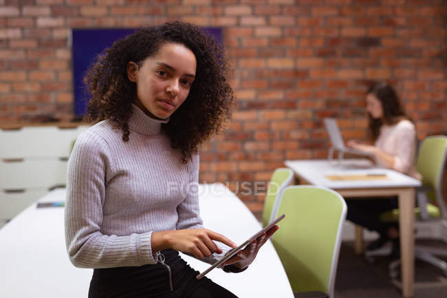 Портрет молодой женщины смешанной расы, работающей в офисе креативного бизнеса, смотрящей в камеру, сидящей на столе с помощью планшетного компьютера, с коллегой, работающей на заднем плане — стоковое фото