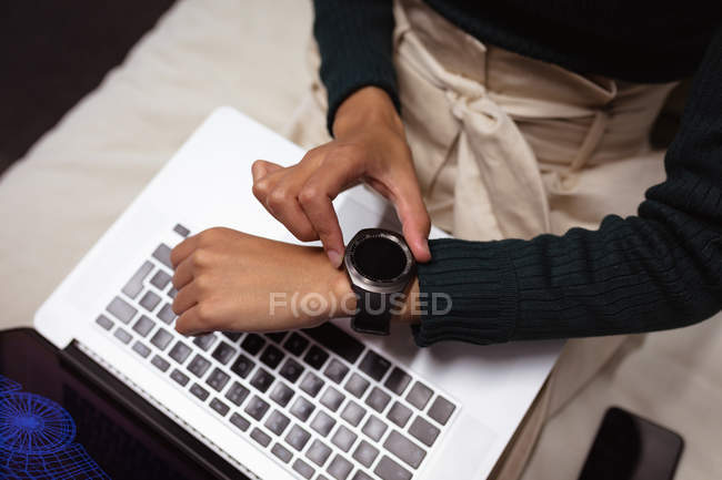 Вид спереду середня секція жінки, яка перевіряє свій смарт-годинник і використовує ноутбук в офісі творчого бізнесу — стокове фото