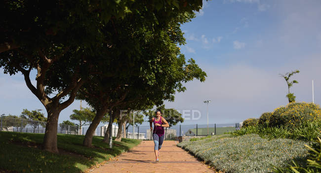 Vue de face d'une jeune femme caucasienne portant des vêtements de sport courant sur un chemin et regardant sa montre intelligente pendant une séance d'entraînement par une journée ensoleillée dans un parc — Photo de stock