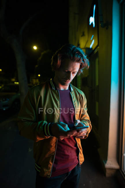 Вид спереду крупним планом молодого Кавказького чоловіка, стоячи на вулиці вночі за допомогою смартфона на освітленому вікні магазину — стокове фото