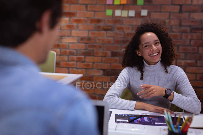 Por encima de la vista del hombro de una joven mestiza y un joven colega caucásico hablando en la oficina de un negocio creativo, la mujer está sonriendo y señalando - foto de stock