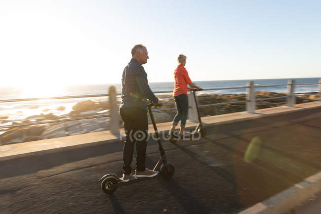 Side view of a mature Caucasian man and woman riding e scooters by the sea at sunset — Stock Photo