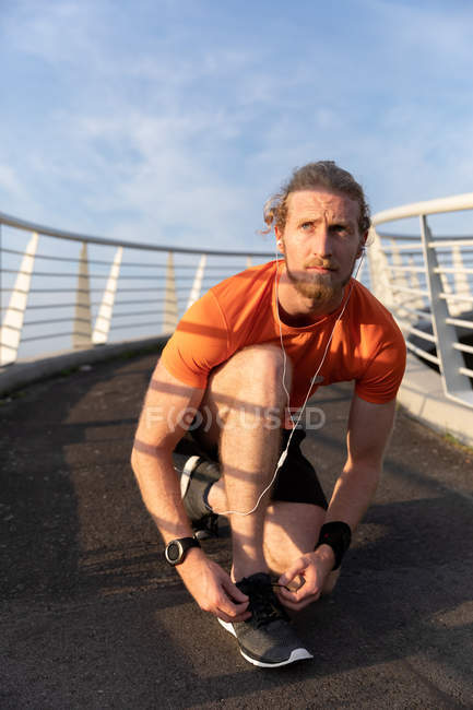 Front view of a young athletic Caucasian man exercising on a footbridge in a city, tying up his laces and listening to music with earphones on during a break — Stock Photo