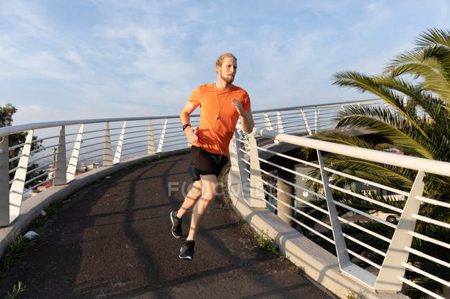 Front view of a young athletic Caucasian man exercising on a footbridge in a city, running and listening to music with earphones on — Stock Photo