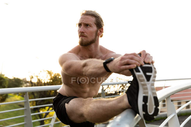 Vista frontale di un giovane atletico caucasico senza maglietta che si allena su un ponte pedonale in una città, che si estende con una gamba sul corrimano — Foto stock