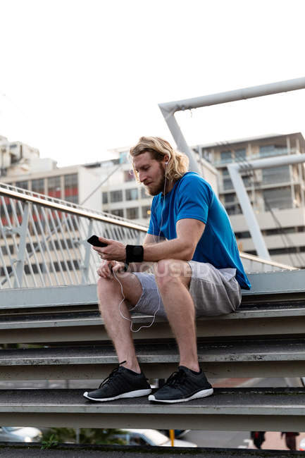Вид сбоку на молодого спортсмена-кавказца, тренирующегося на пешеходном мосту в городе, сидящего на ступеньках и пользующегося смартфоном во время перерыва — стоковое фото