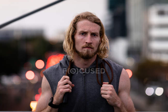 Portrait of a young athletic Caucasian man exercising in a city park in the evening, resting during a break with a skipping rope round his neck with defocused city lights in the background — Stock Photo