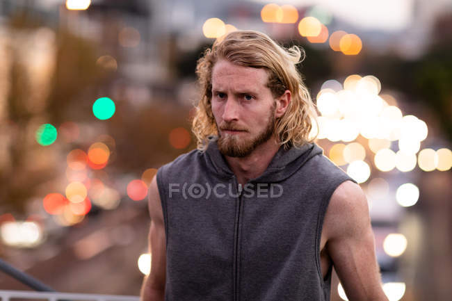 Retrato de um jovem atlético caucasiano exercitando-se em um parque da cidade à noite, olhando para a câmera durante uma pausa com luzes da cidade desfocadas no fundo — Fotografia de Stock