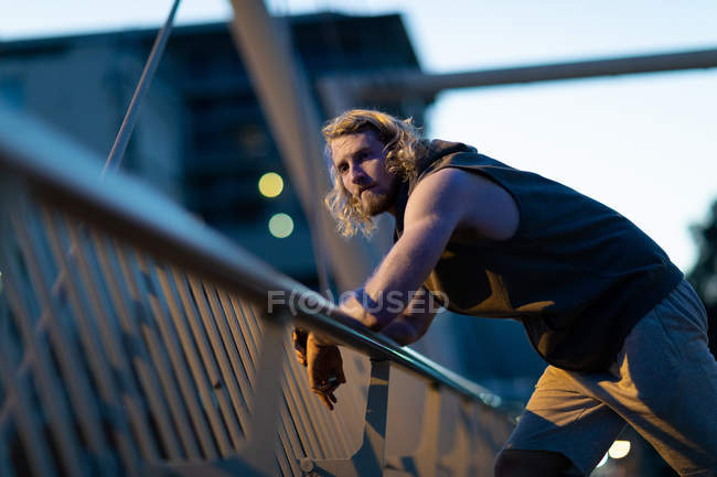 Portrait of a young athletic Caucasian man exercising in a city park in the evening, resting on a handrail during a break with defocused lights in the background — Stock Photo