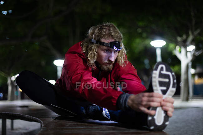 Vista frontal de um jovem atlético caucasiano exercitando-se em um parque da cidade à noite, estendendo-se em um banco com um farol ligado — Fotografia de Stock