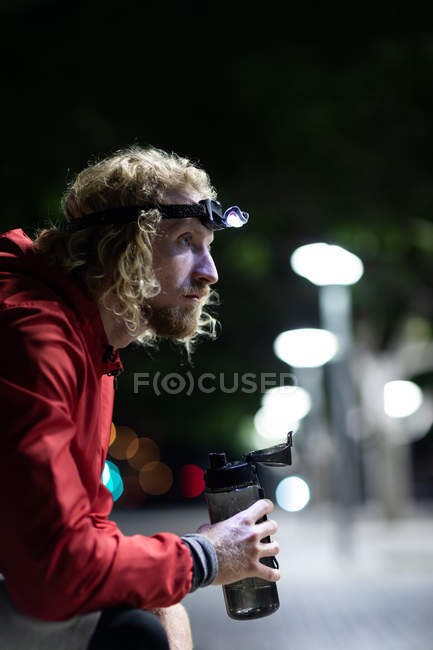 Side view of a young athletic Caucasian man exercising in a city park in the evening, with a headlight on resting during a break with defocused city lights in the background — Stock Photo
